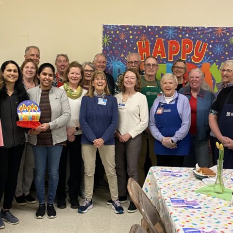 A group of men and women in front of a Happy Birthday sign. In the foreground is a brightly decorated table, ready for guests to come in.