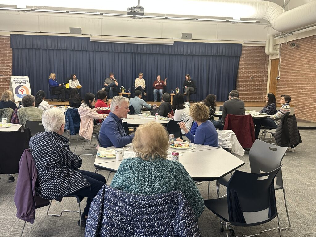 People sit at octagonal tables facing a number of people who are seated on stage.