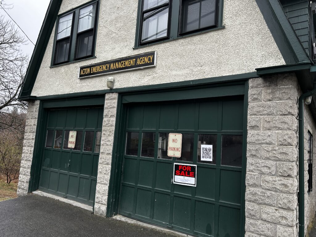 An old stone building with green garage doors and a For Sale sign. The sign over the doors says "Acton Emergency Management Agency."