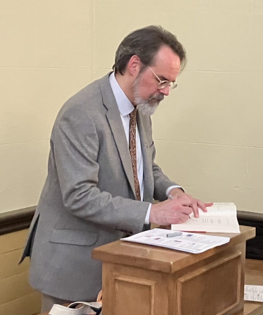 A bearded man in a suit signs a book at the podium.