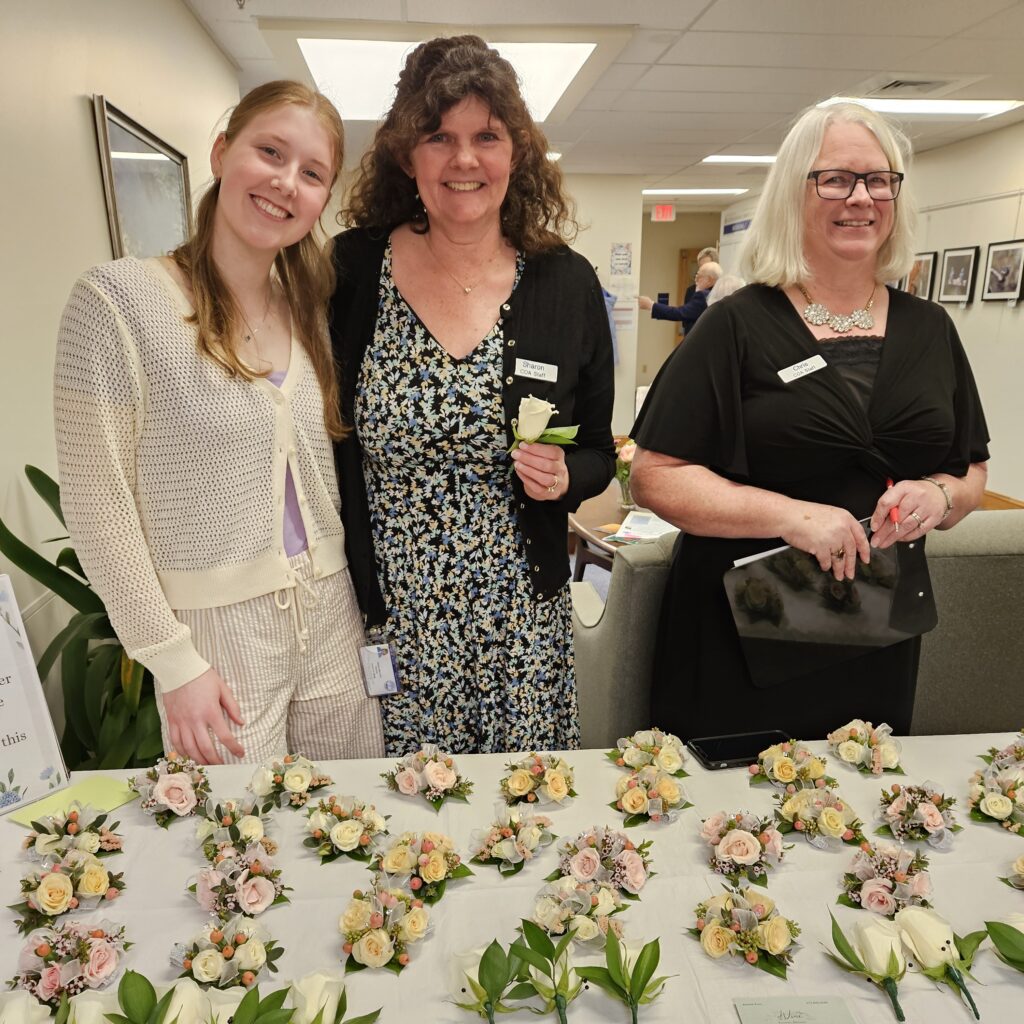 A young woman and two older women smile in front of a table full of beautiful corsages and boutonnieres. The boutonnieres are all white roses and the corsages are in pink and apricot.