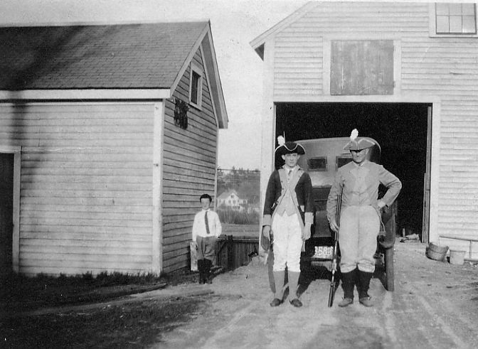 A black and white photo. On the left, a young boy wearing a white shirt, tie, and knickers stands by the garage. In the center of the picture, a young man and his father, wearing colonial garb stand in front of a car, perhaps a Model T? The older man has his musket in hand.