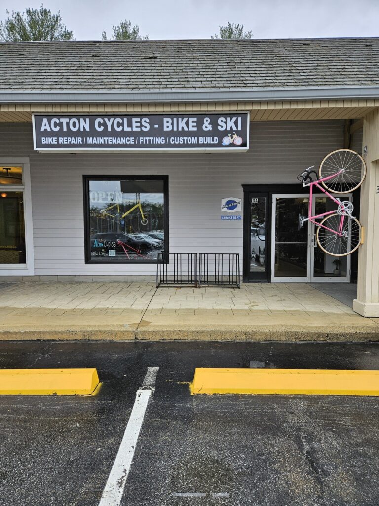 The Acton Cycles - Bike & Ski sign. A bike rack is in the front of the building and a pink bike hands vertically from a post in front of the building.