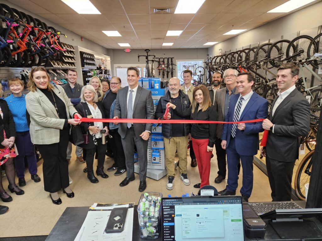 A group of people in a shop with bicycles on the wall. A balding man (the owner) cuts the ribbon with oversized scissors.