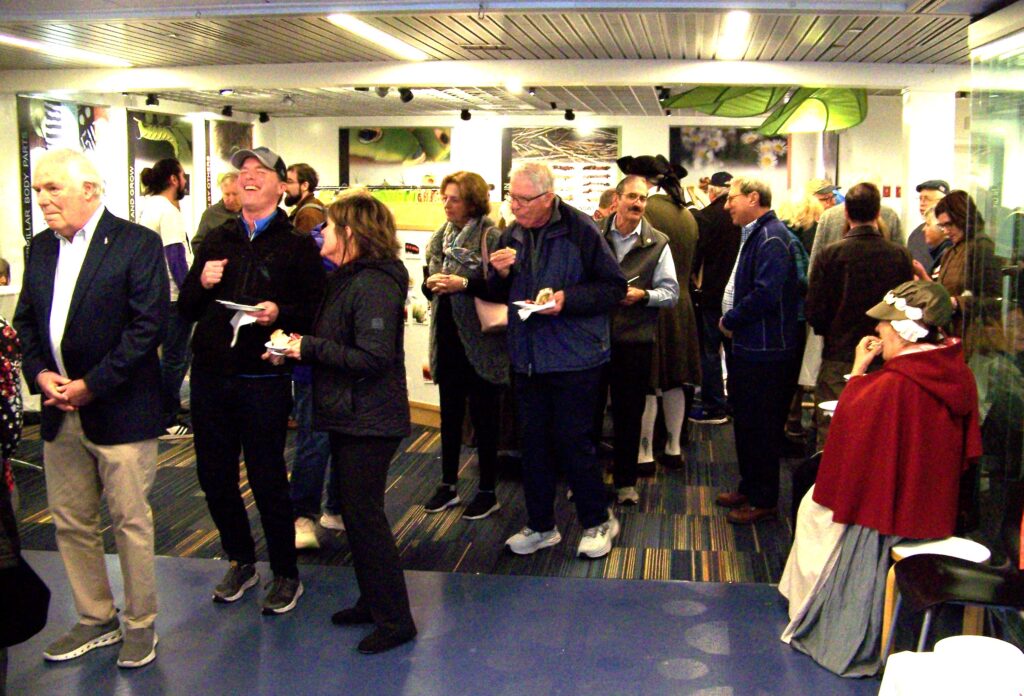 People laughing, talking, and eating while standing in a line. Most people are dressed for this century, but people in Colonial garb are also around.