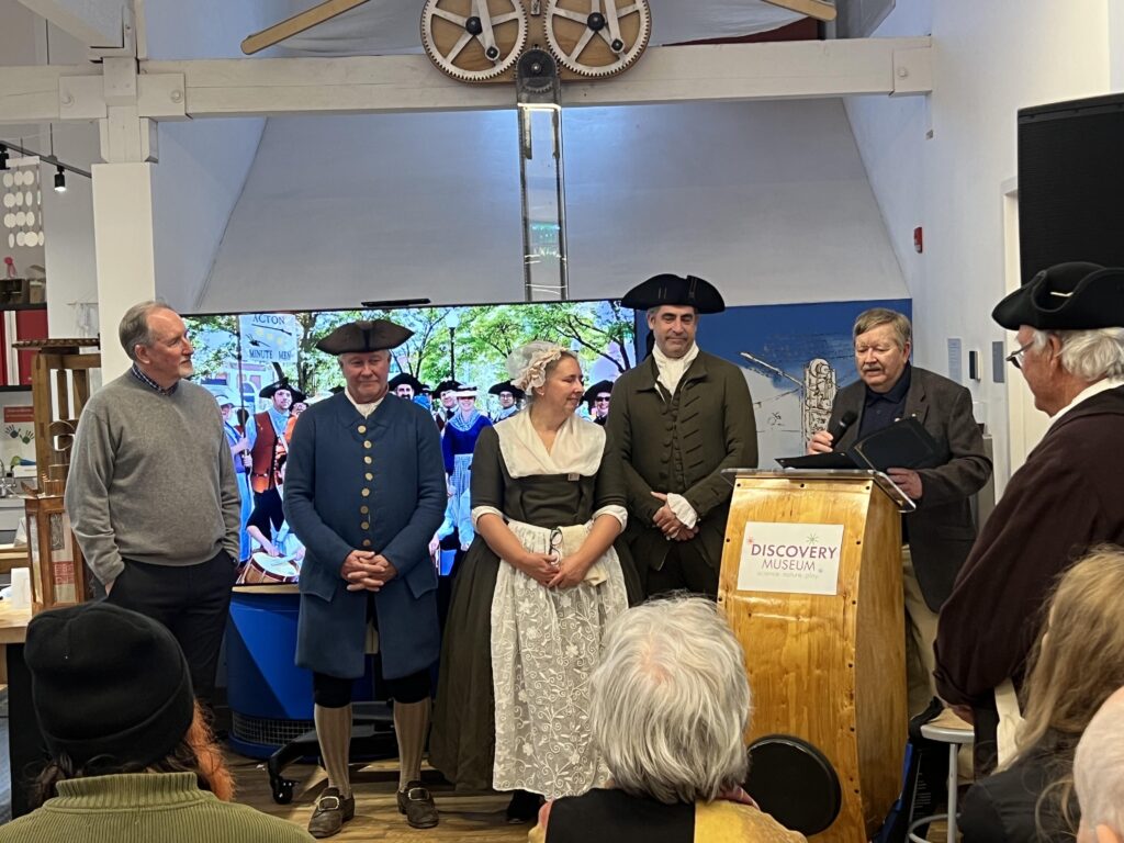 A man stands at podium. To his left, three people in Colonial garb and one man wearing a sweater listen intently.