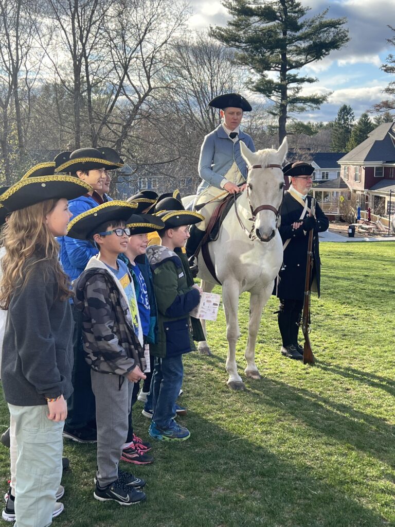 A woman wearing Revolutionary War men's clothing is sitting astride a white horse. They are on a lawn and a group of kids wearing tri-corner hats stands next to her. On the other side, a fully kitted Minuteman (including musket) stands by.