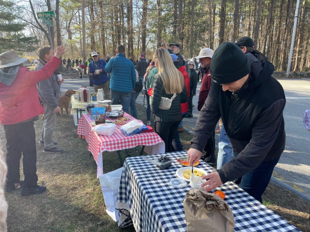 Several tables filled with food, coffee, and napkins are ready for the marchers. In the foreground, a man spreads cream cheese on a bagel. Neighbors mill around and you can see the marchers in the background.