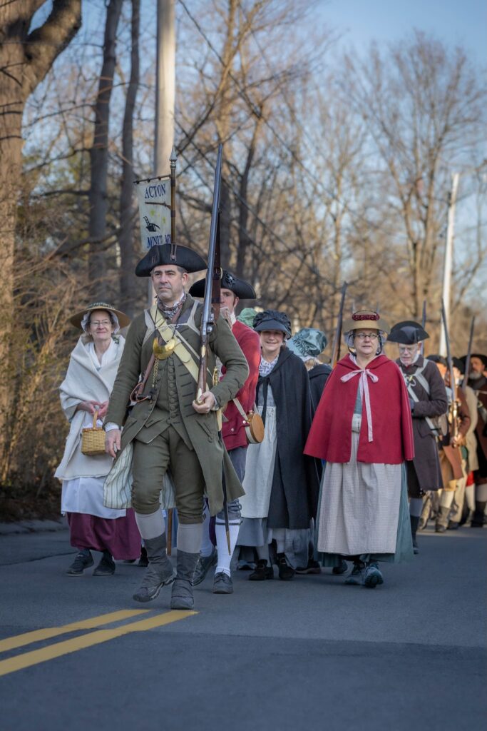 A group of people in colonial garb walk down a road in the early morning light.