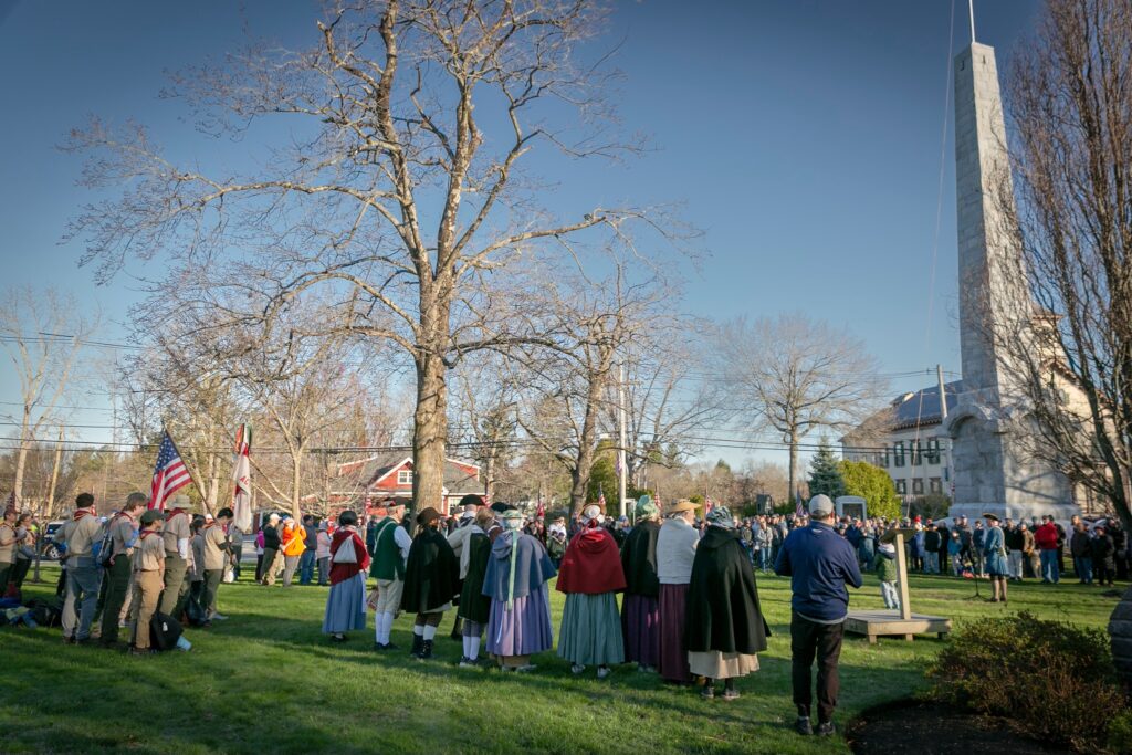 From the distance, Minuteman Steve Crosby is explaining the history of April 19, 11775. Acton's monument is in the background and there are several hundred people listening.