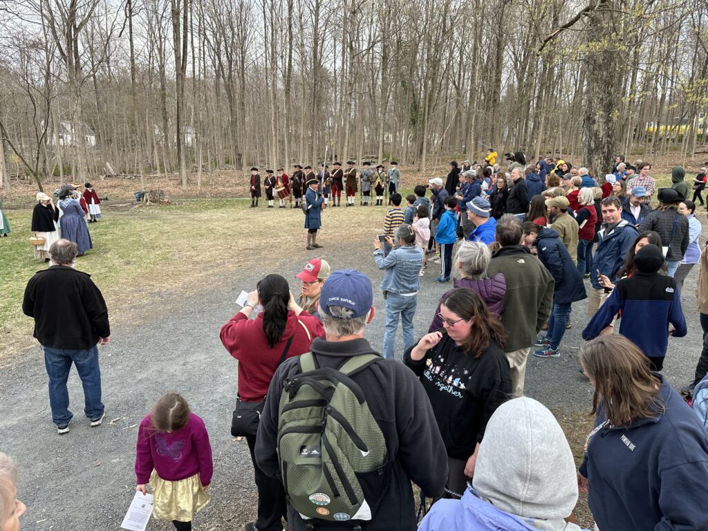 A large group of people watch a man in colnial garb give his final speech of the day. In the background are the re-enactors from the Acton Minutemen and Ironwork Farm.