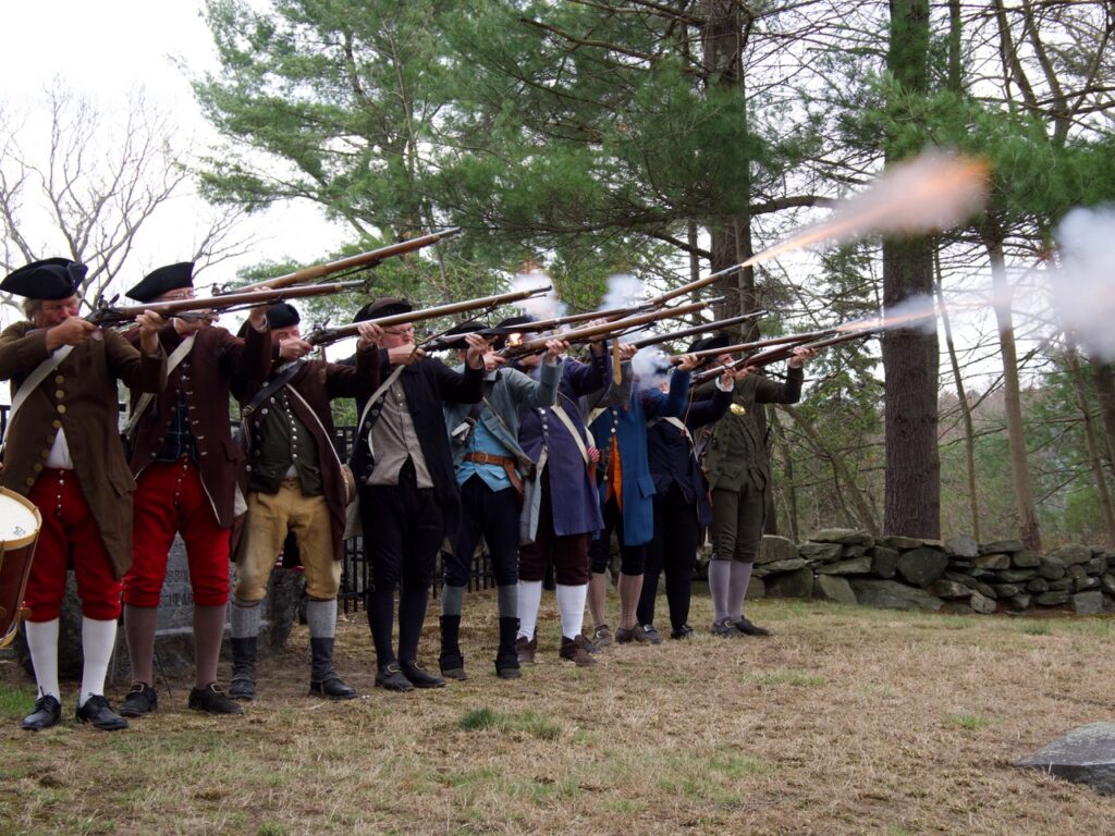 A group of men and women in men's colonial garb shoot their muskets into the air. Smoke and fire is visible from some of the guns.