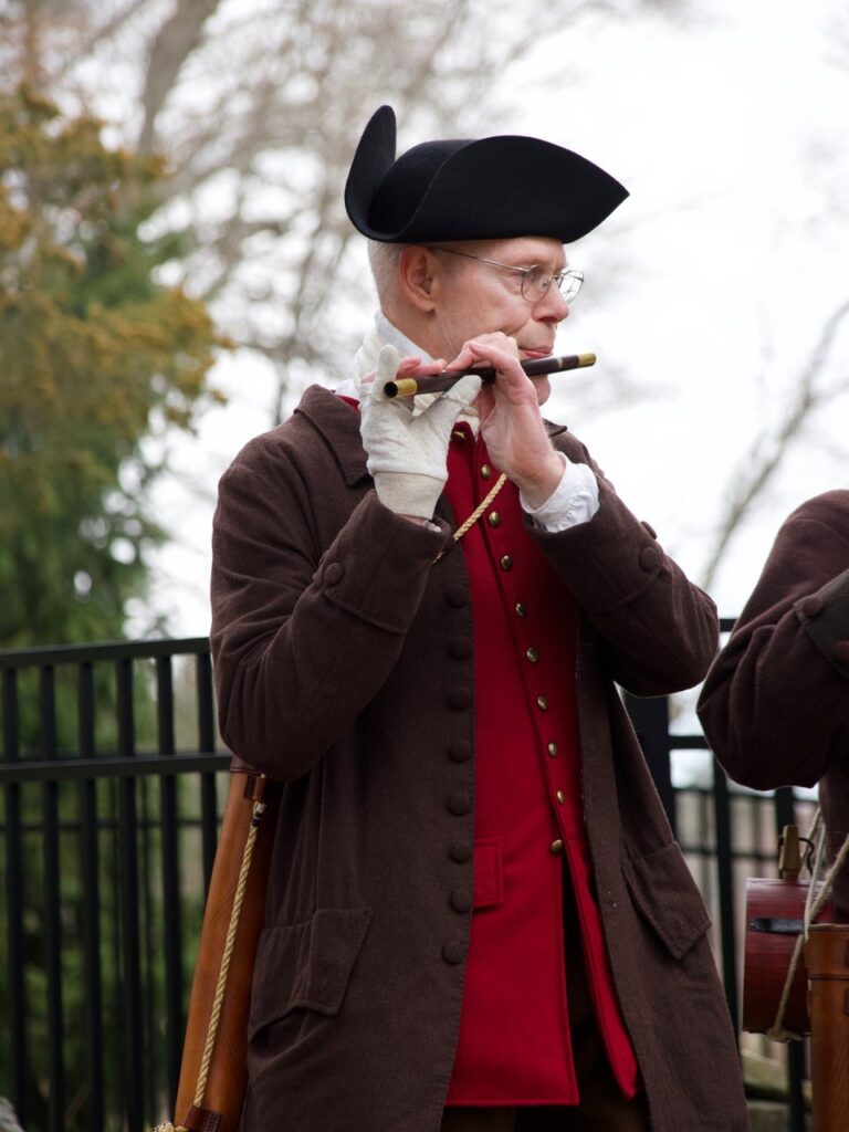 A tall man wearing a tricorner hat, brown jacket and red vest playes a fife.