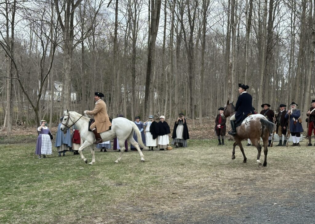 The riders and their horse make it up the hill to the yard in front of Faulkner house. Minutemen and women stand in the background.
