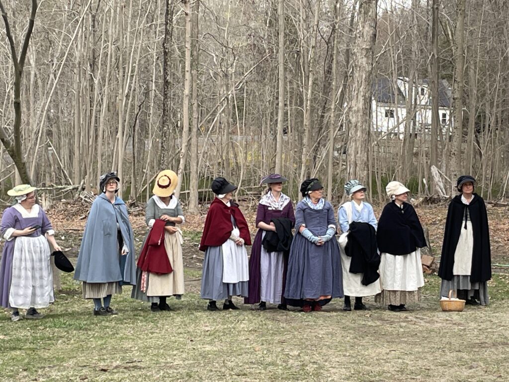 A large group of women wearing colonial garb (long dresses, aprons, bonnets, and capes) stand in front of woods.
