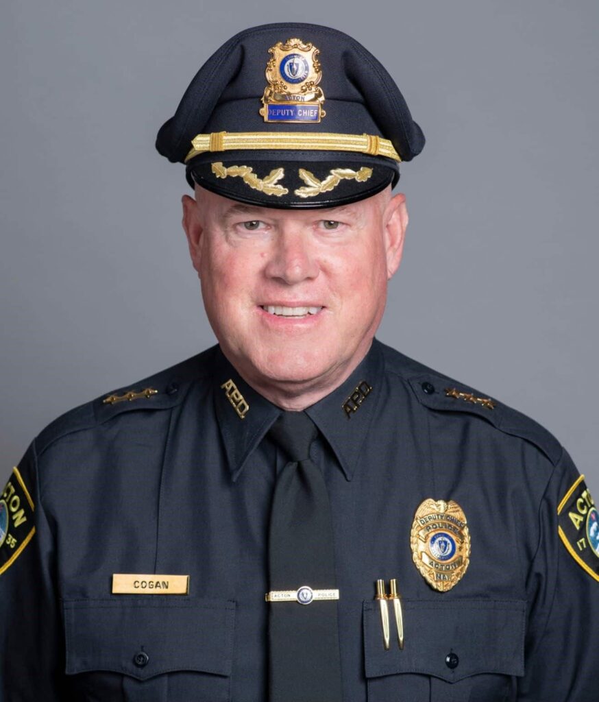 Formal portrait photograph of a mature, white man, wearing a dark blue uniform shirt, tie and hat. Nameplate says "COGAN." Shoulder patches say "ACTON." Gold-colored badge says "Deputy Chief, Police, Acton, MA."