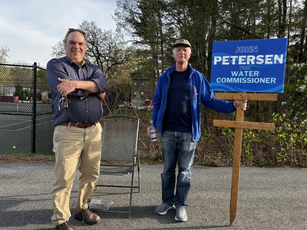 Two men stand at the corner of the AB tennis courts. The man on the right holds a John Petersen for Water Commissioner sign.