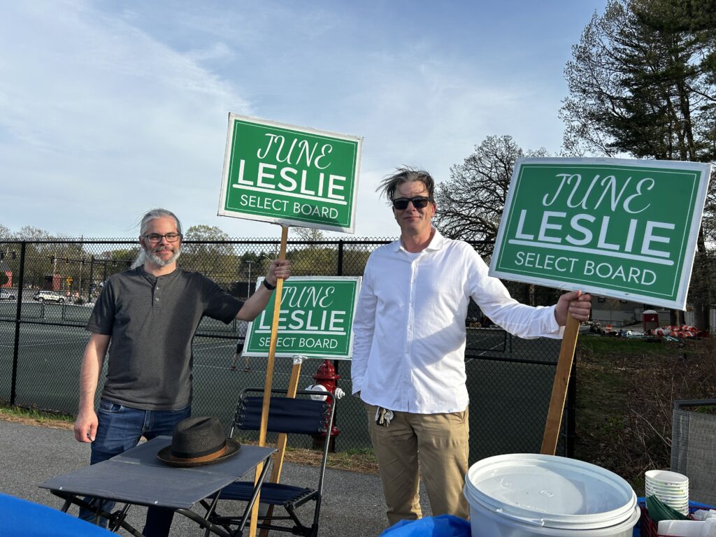 Two men, one with a beard and ponytail, hold green June Leslie signs. A table and chair is set up to allow the helpers to take a break.