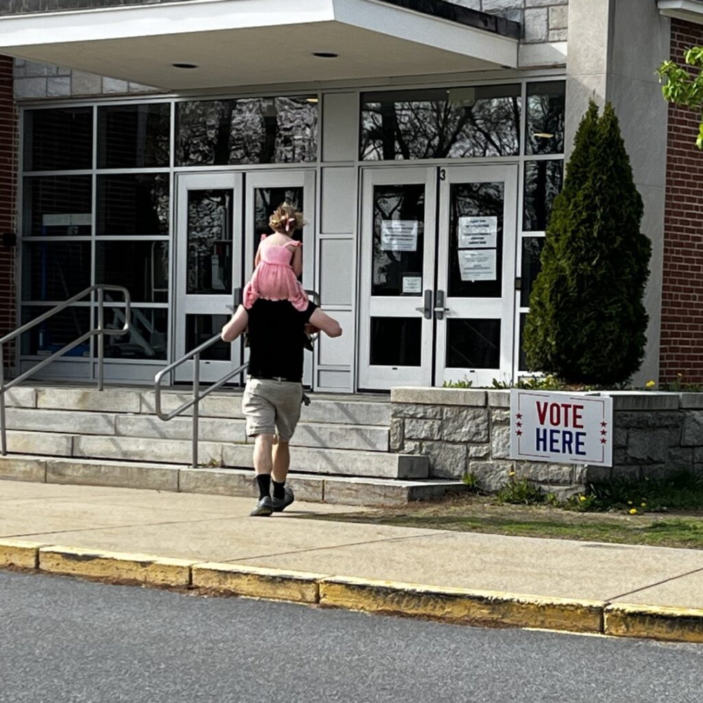 From the back, a man walks up to the steps of the polling location. A girl in a pink dress rides on his shoulders.