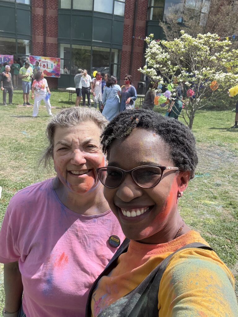 Two women, one white, one Black have smears of dye all over their faces and clothes. There are lots of people in the background.