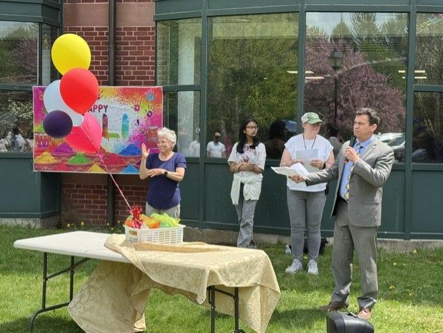 In front of the Library windows, a man in a suit speaks into a microphone. There are a couple of other people in the background, but on the left, a woman in a blue T shirt is signing his speech. Brightly colored balloons and a Happy Holi sign are part of the scene.