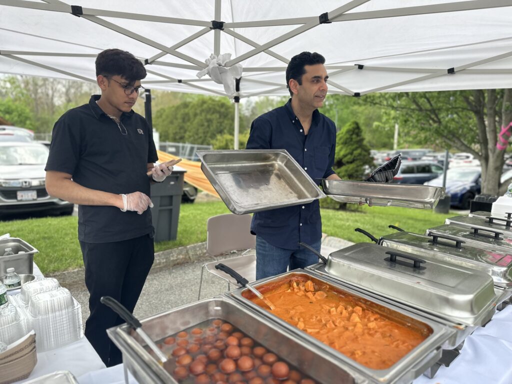 Two men stand behind catering trays of chicken tikka and galub jamun,