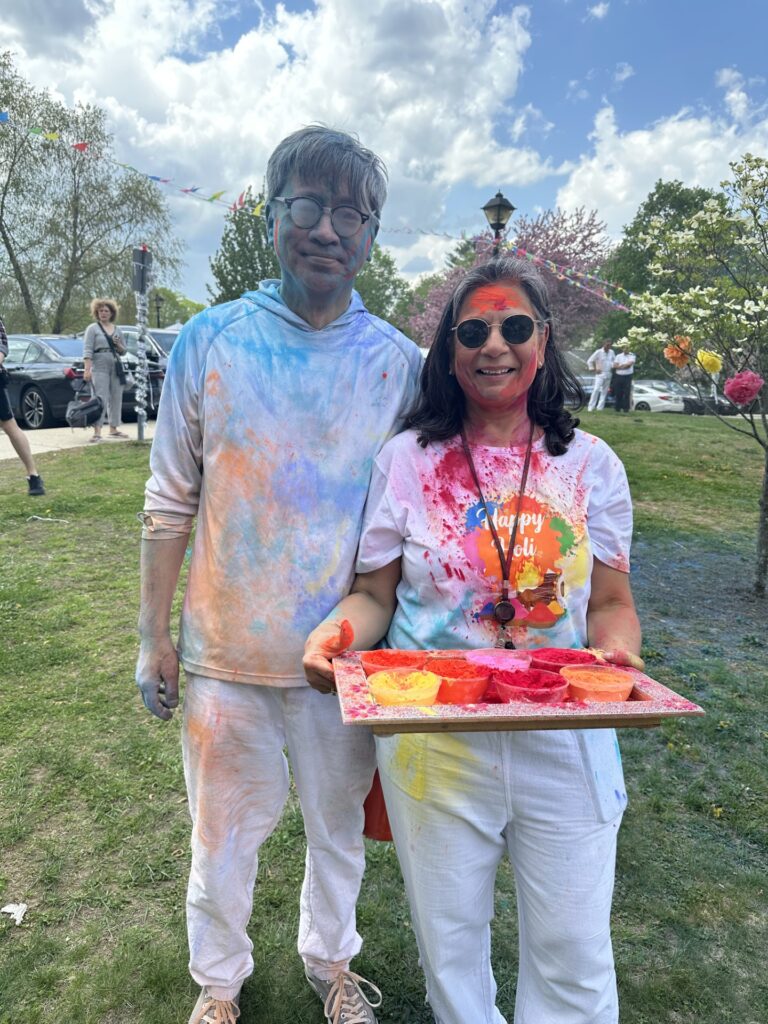 A man and woman stand in the sun. They are both wearing white and covered with Holi powder. The man's eyeglasses are coated and he's smiling slyly. The woman is carrying a tray of dye powders.