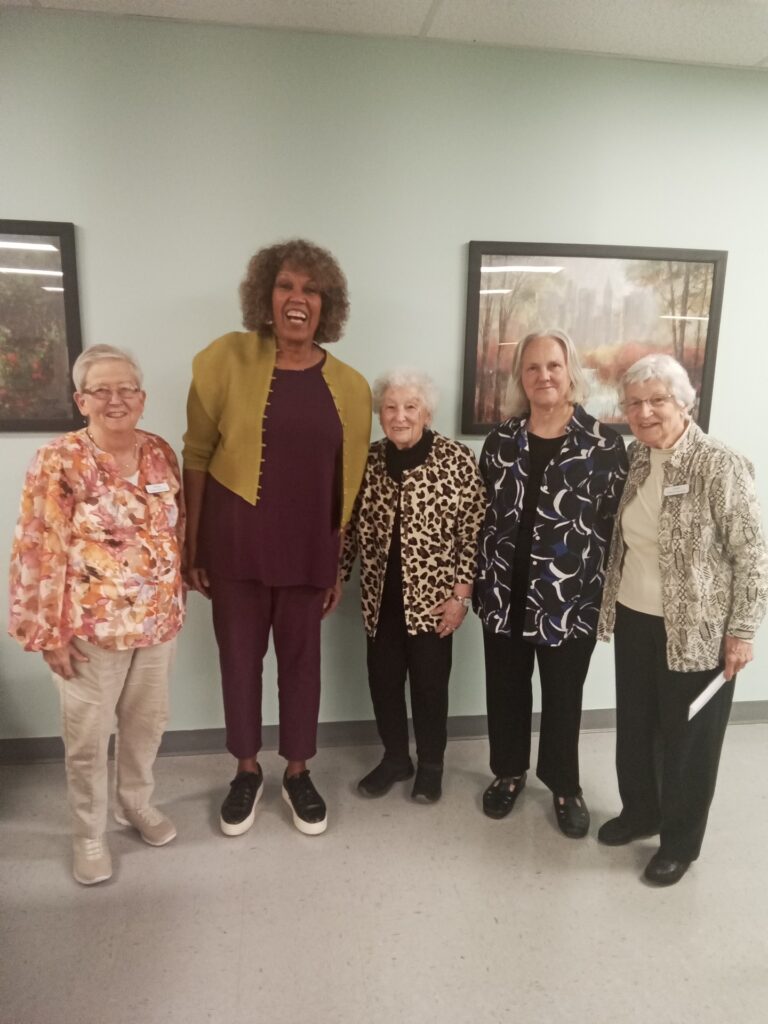 A very tall smiling woman with curly hair towers over four older grey-haired women.