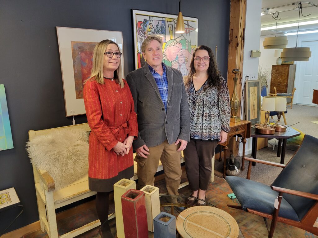 A woman in a red dress, a man in a sport jacket, and a woman in a brown and white tunic and slacks stand in front of a bench with a furry pillow. There are two paintings in the background and interesting items all around.