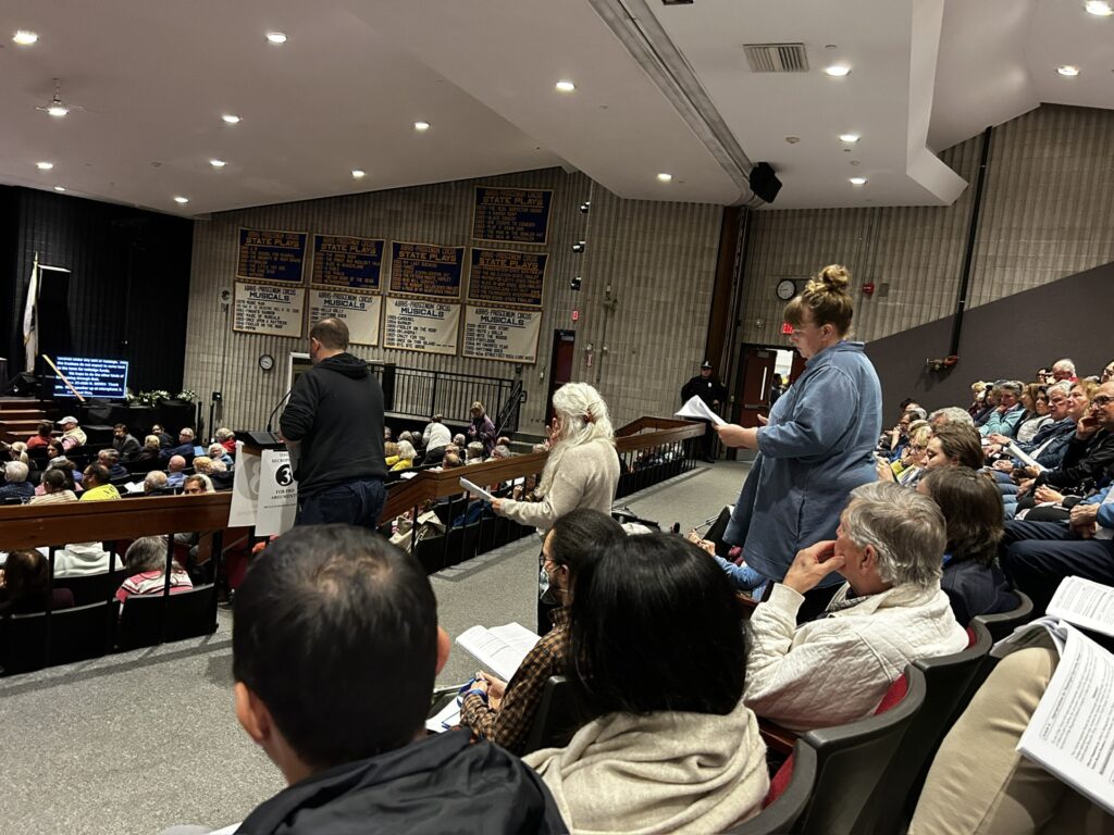 One man speaks at a microphone and two women are in line behind him. Many people are seated in the auditorium.