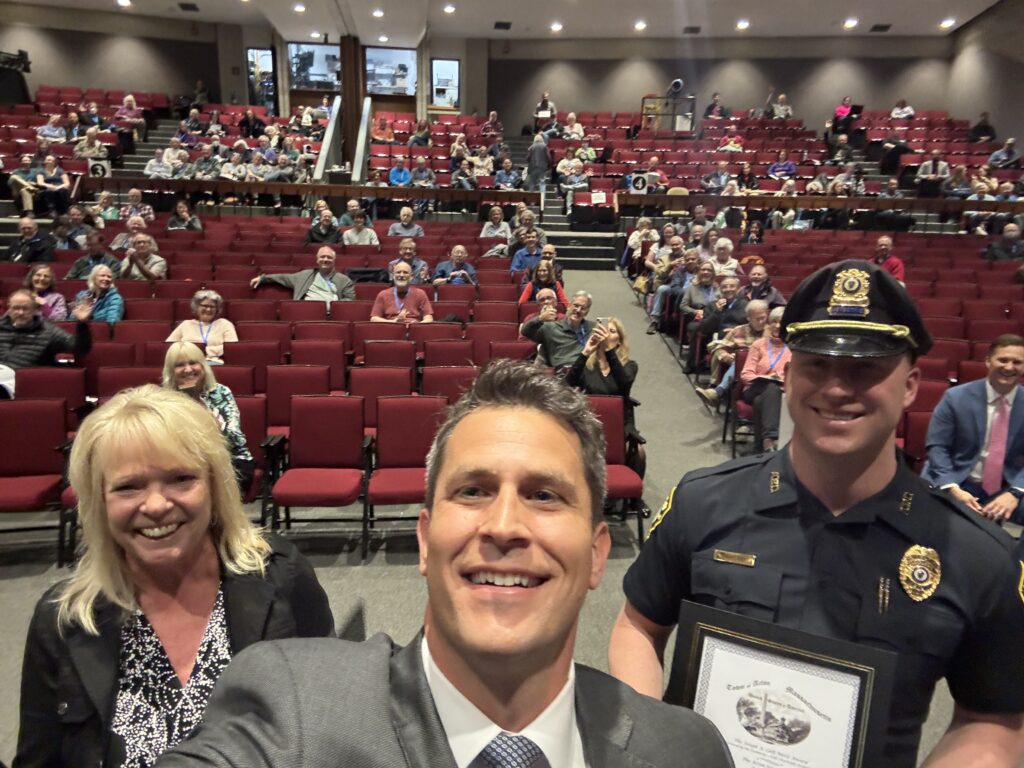 A blonde woman, a man in a suit, and a tall man in a police uniform and are in the foreground. The mostly empty auditorium is in the background.