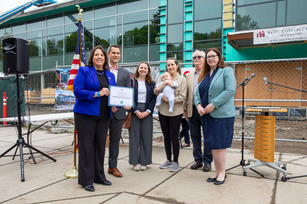 Six people stand in front of a building that is still under construction. One smiling woman holds a very cute baby. The woman on the far left (Lt Gov Driscoll) is standing next to Town Manager John Mangiaratti. They are smiling and holding a citation.