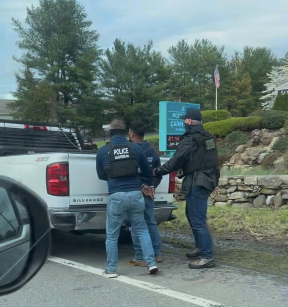Two men wearing Police vests and face coverings handcuff a third man against a white truck.