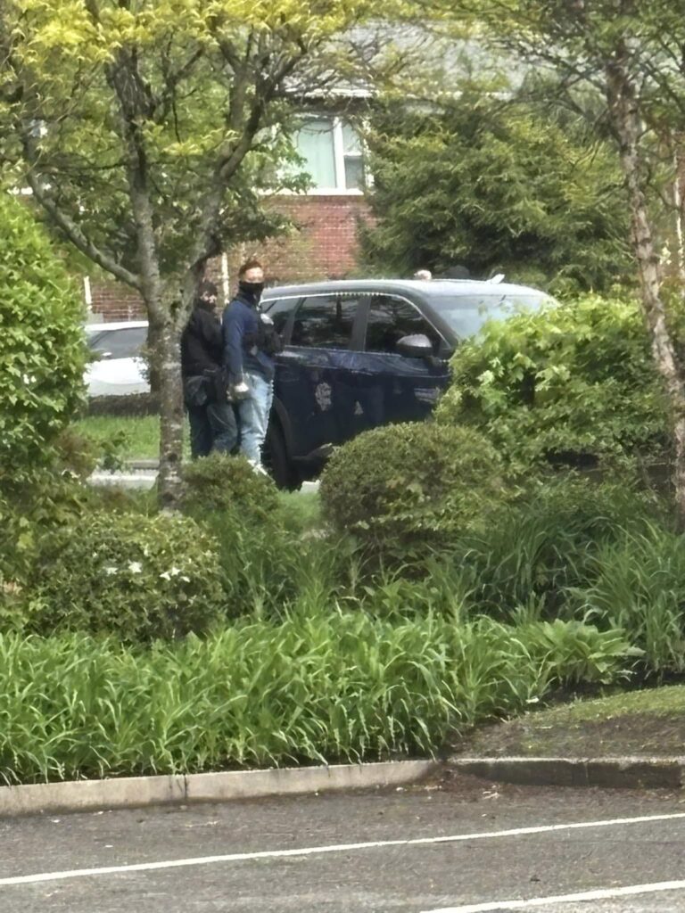 Landscaping foliage dominates the perimeter of the photograph, with a red brick building in the background. A parked black car is visible behind foliage. Next to the car, stand two people, with black masks or high collars obscuring the lower parts of their faces.