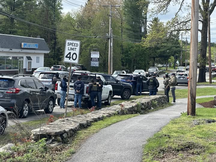 Multiple vehicles parked haphazardly along side of the road. A cluster of people next to a white pickup truck includes one person with arms apparently shackled behind them, and others with black face mask.