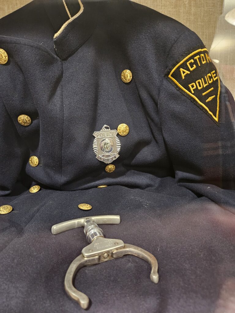 Photograph of a historic police uniform jacket, with gold buttons, badge saying "Police," and shoulder patch saying "Acton Police." An old-fashion hand cuff is lying on top of the jacket.