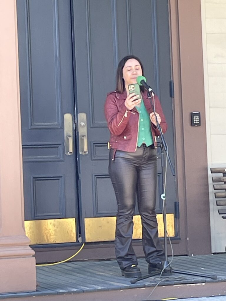 A woman wearing a leather jacket and pants stands in front of a microphone reading from her phone. A black door is in the background.