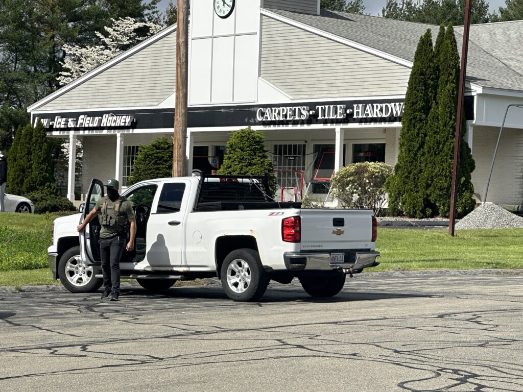 Man with tactical vest and black face mask emerging from white pickup truck.