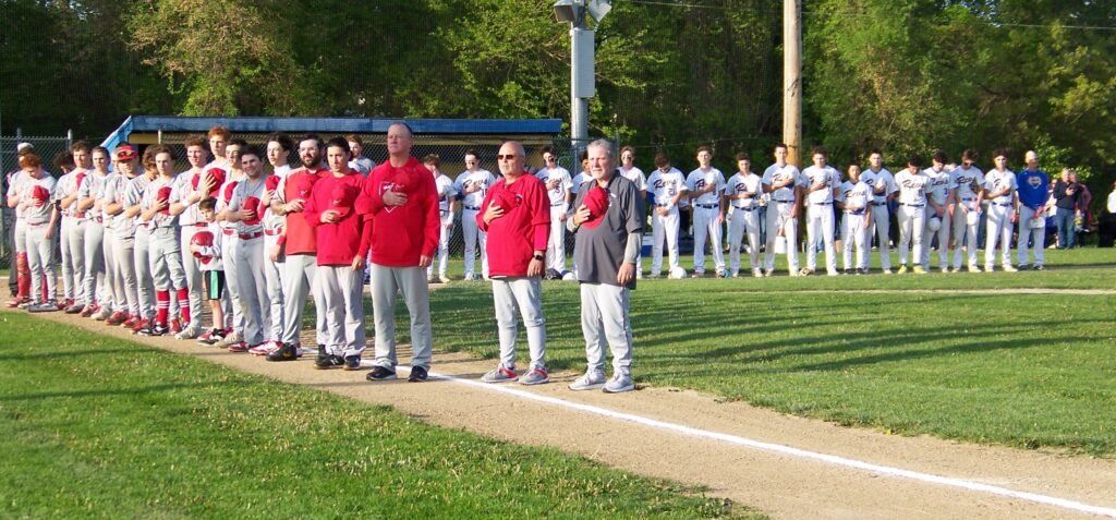 Players and coaches stand in a line with their caps over their chests.