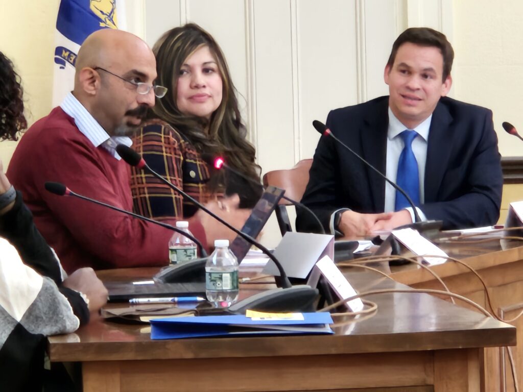 A man in a red sweater speaks into a mic. A woman and man listen intently.