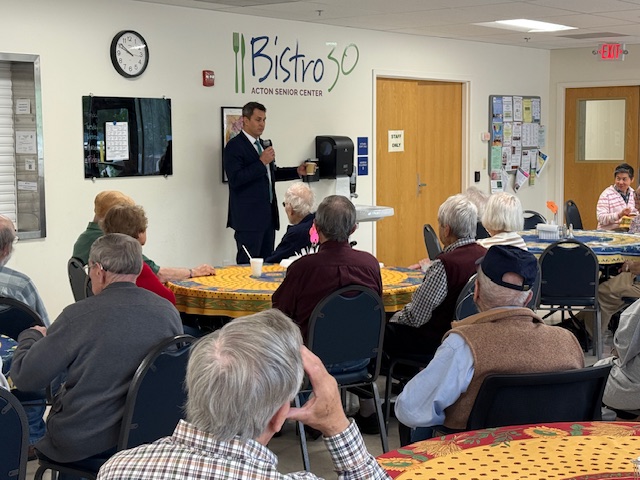 A tall man in a suit speaks to an audience in front of the Bistro 30 sign at the Acton Senior Center.