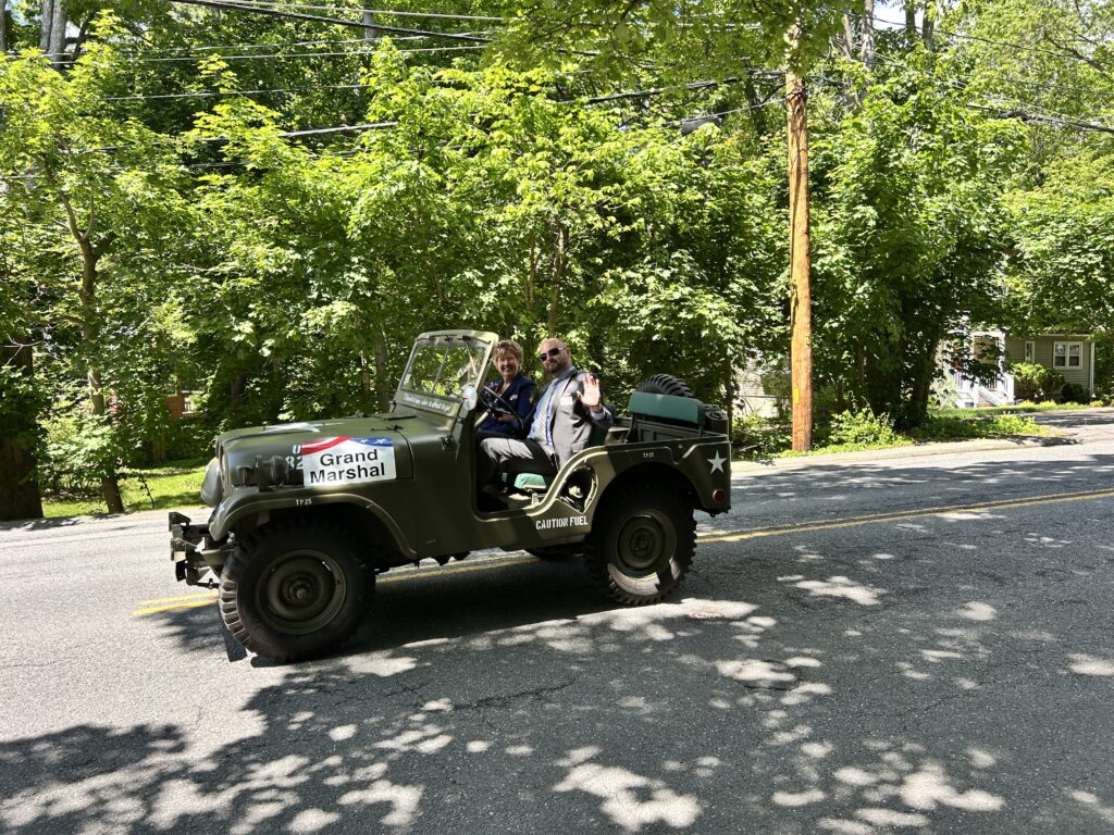 Two people ride in a green Jeep with a Grand Marshal sign. The male driver is waving.