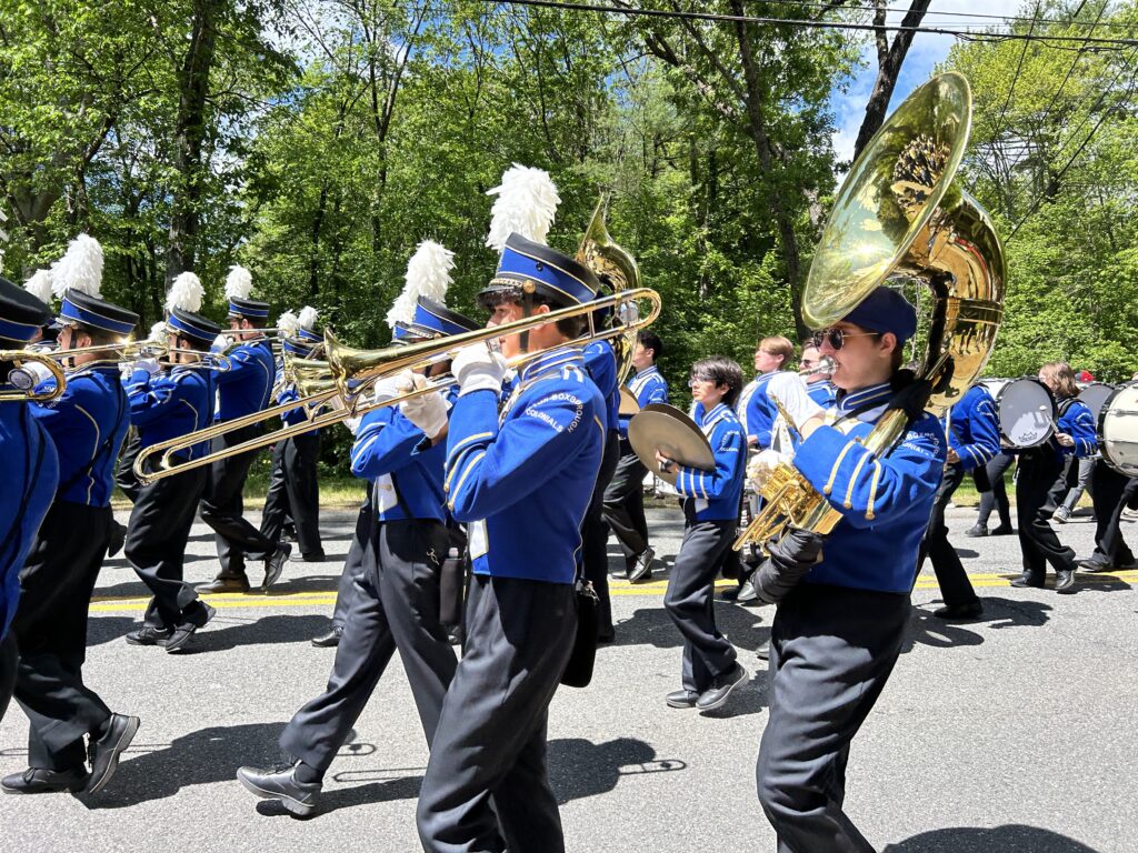 A group of students in blue and black uniforms play brass instruments. Most of the marchers are wearing plumed hats for added oomph.