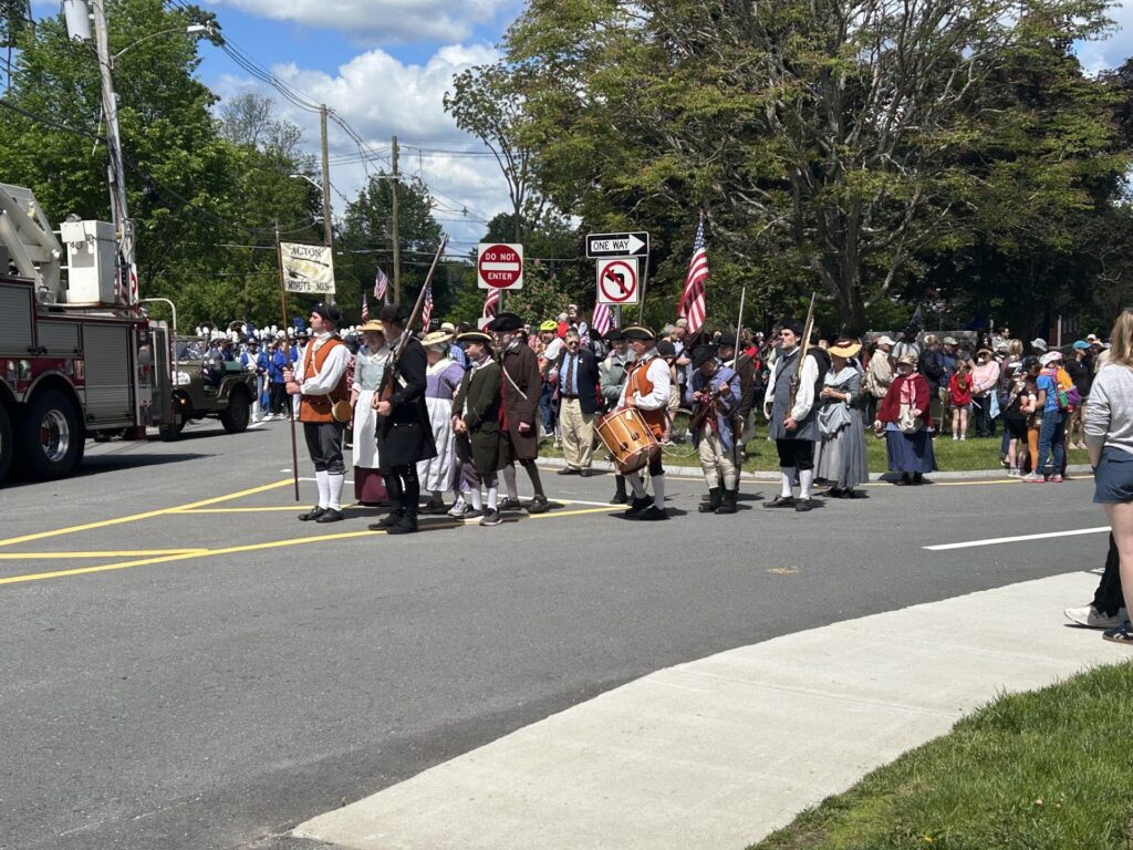 A group of men and women in Colonial dress line up at the corner of Main and Concord Road. the band and other marchers are in the background.