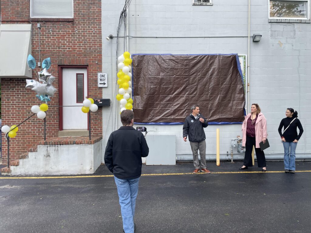 A building wall. The mural is hidden behind a brown tarp, but there are yellow and white balloons along the side.