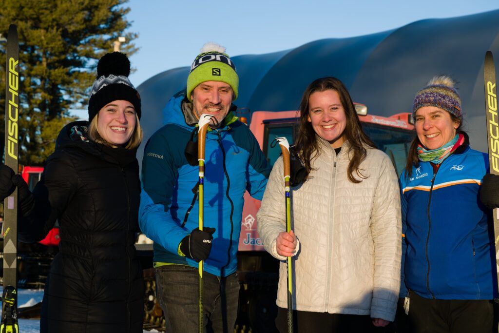 Three women and a man wearing winter coats and holding skiis and poles smile at the camera.