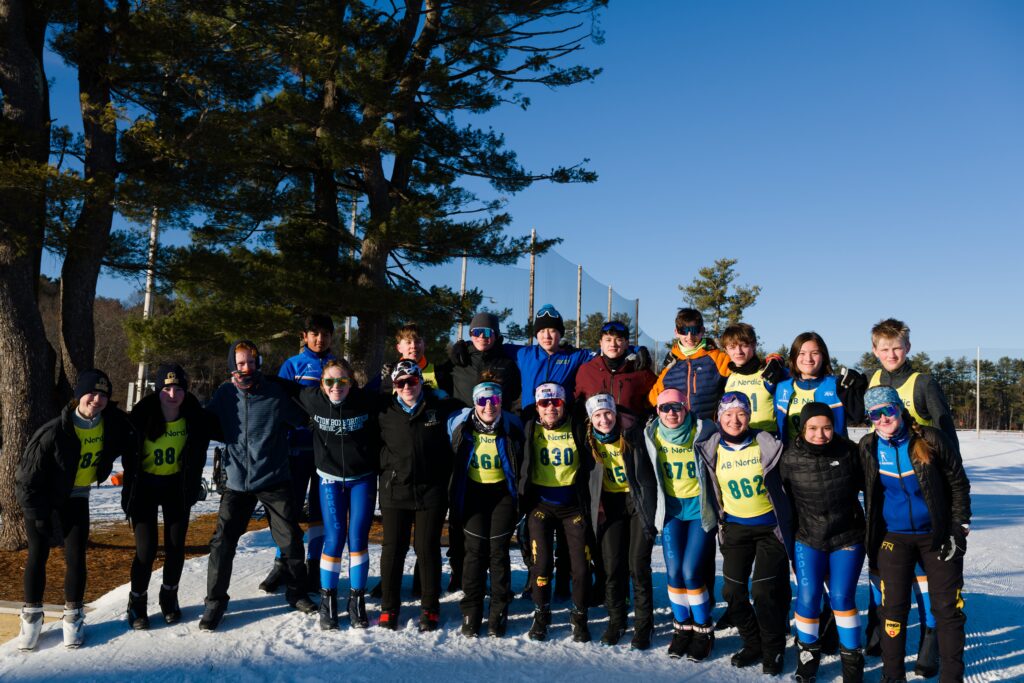 A group photo of teens and adults on a sunny day in the snow. All of the kids are wearing Nordic ski gear, and some are wearing racing bibs.