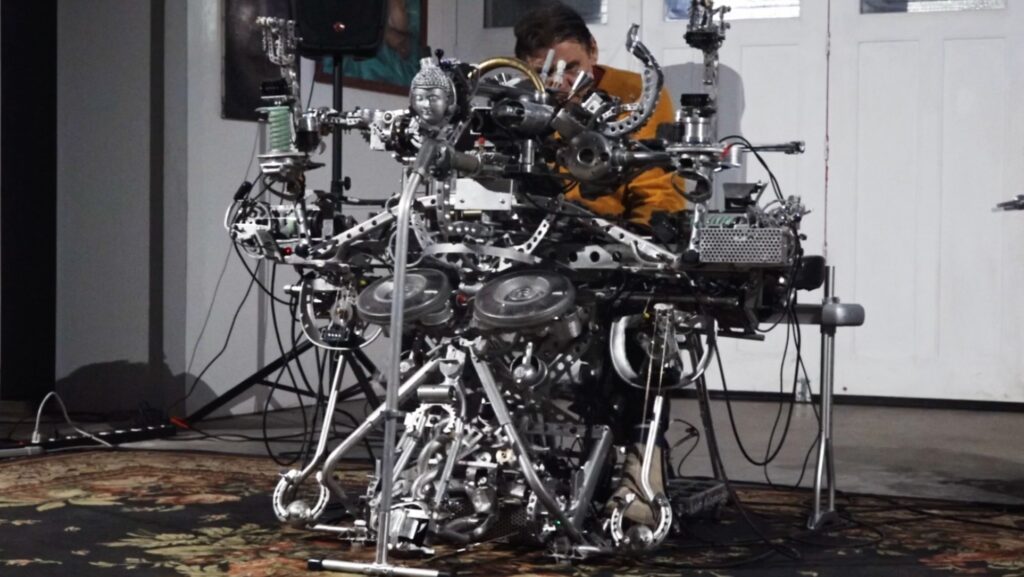 A man sits behind a contraption of metal parts. The instrument looks less like a musical instrument and more like a robot gone wild.