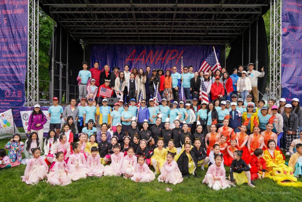 A large group of people stand on an outdoor stage and spill out onto the lawn. Many are holding American flags and there are groups of children in bright costumes.