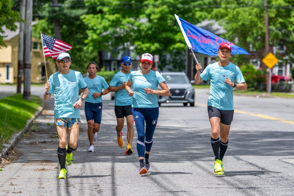 A group of runners wearing blue t-shirts on a sunny day. The runner on the left is carrying an American flag. The runner on the right is carrying a blue flag with red writing.